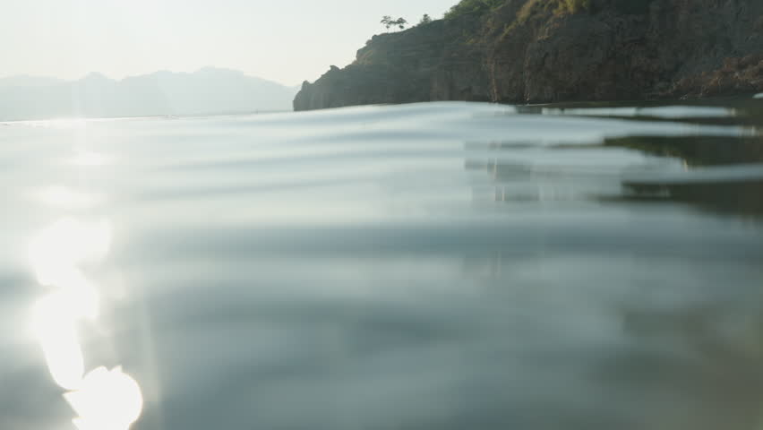The Camera Descends Below the Water Surface, Transitioning from a Sunlit Coastal Landscape to Mysterious Dark Underwater Rocks. Slow motion.