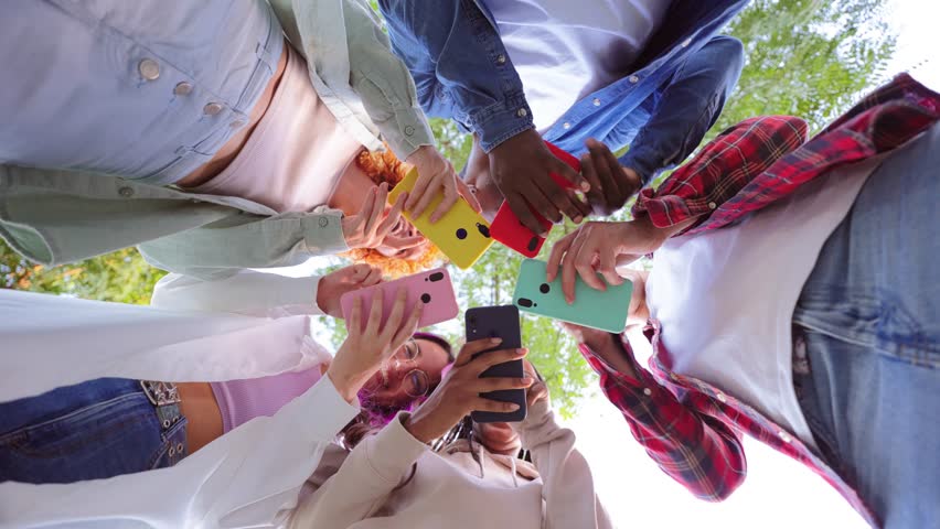 Low angle view of group of friends standing in circle, all using cellphones outdoors. Vibrant smartphone colors contrast the sky, showing modern technology and social connection in urban setting. High