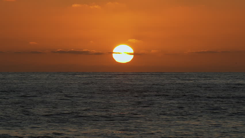 Golden orange sun sets through thin cloud over calm dusk ocean water