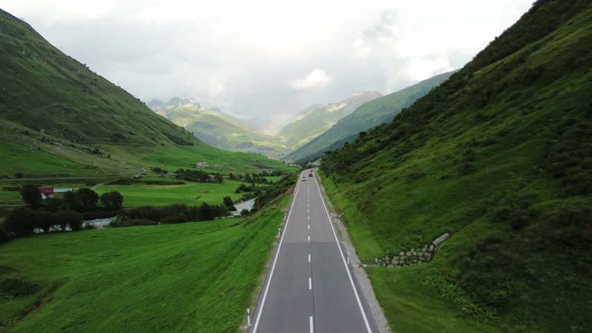 Road in Swiss Alps with one car and rainbow at the sky shot from a drone