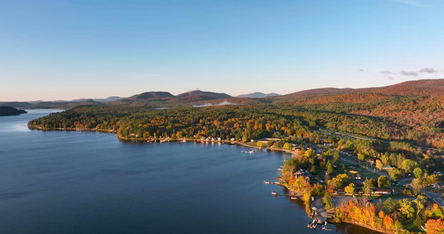 A stunning sunrise over Schroon Lake, NY, with warm light casting a soft glow on the calm water and surrounding autumn-colored forests and mountains.