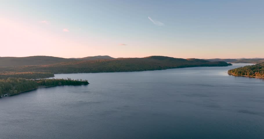 A stunning sunrise over Schroon Lake, NY, with warm light casting a soft glow on the calm water and surrounding autumn-colored forests and mountains.