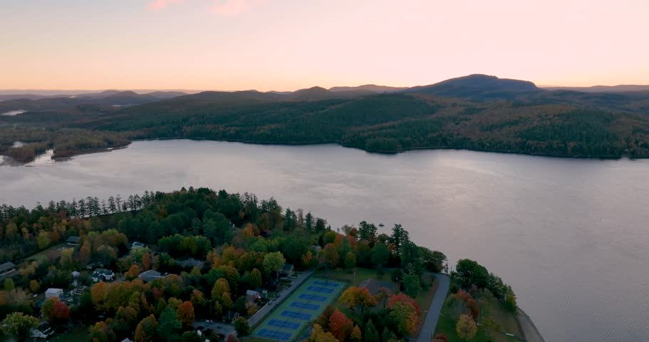 A stunning sunrise over Schroon Lake, NY, with warm light casting a soft glow on the calm water and surrounding autumn-colored forests and mountains.