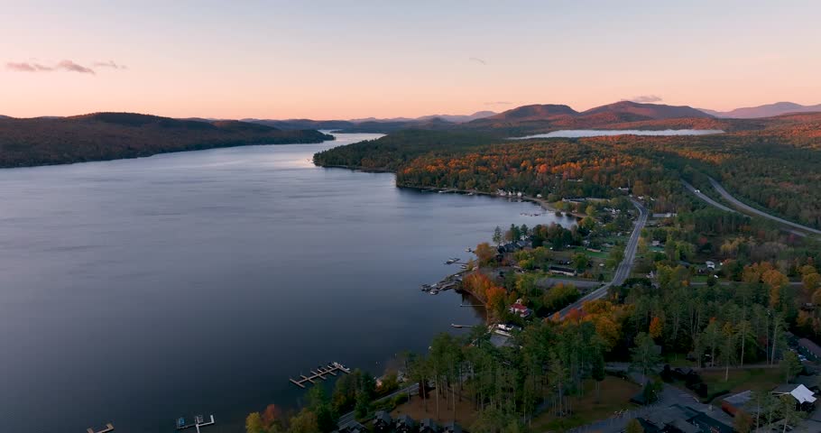A stunning sunrise over Schroon Lake, NY, with warm light casting a soft glow on the calm water and surrounding autumn-colored forests and mountains.