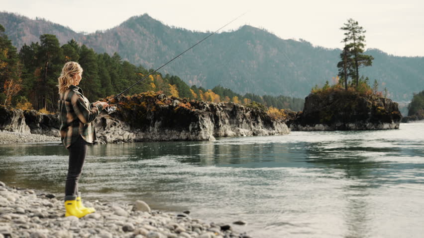 A woman fishes on the rocky river shore surrounded by mountains and autumn forest, enjoying a calm outdoor moment in nature.