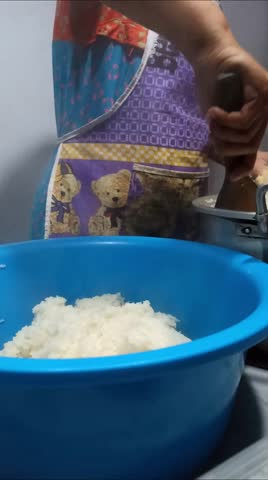 A woman wearing an apron is cooking rice to make nasi uduk, a traditional Indonesian dish made from steamed coconut rice, often served with various side dishes.