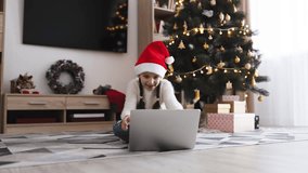 Young Caucasian girl wearing Santa hat sitting on floor engaged in online video call. Christmas tree with decorations and gifts in background creates festive atmosphere. - Powered by Shutterstock - Get 15% off with code: PIKWIZARD15