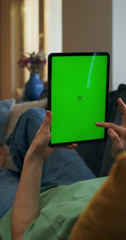 A woman uses a tablet with green screen lying on the couch at home, an unrecognizable person