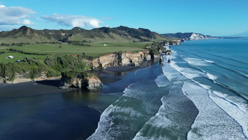 An aerial slow-motion of the sea waves near green rocky shore with cliffs at the Three Sisters Lookout in Tongaporutu, New Zealand