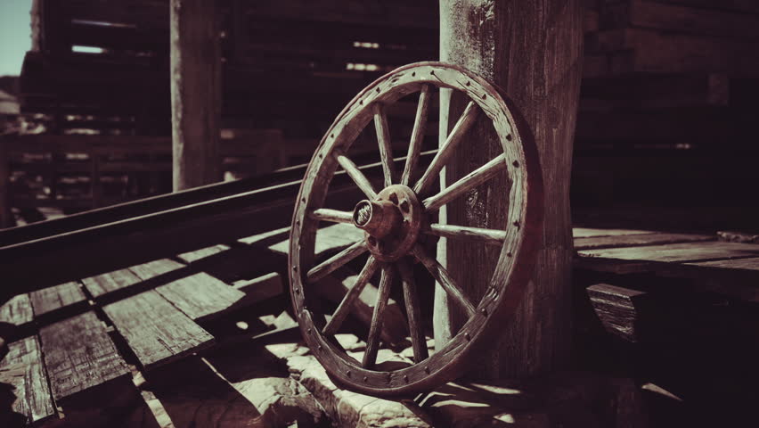 A rustic wooden wagon wheel leans against a weathered post inside an old barn, surrounded by dim light filtering through cracks. The scene evokes a sense of nostalgia and history.