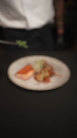 A slow-motion vertical footage of putting herb in a plate contains a piece of Salamon and fried vegetables