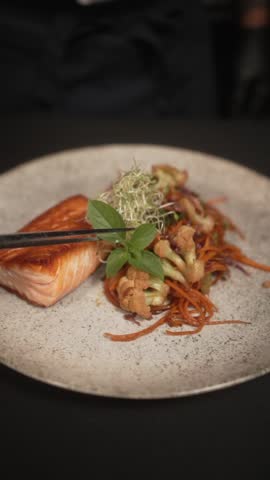 A slow-motion vertical footage of putting herb in a plate contains a piece of Salamon and fried vegetables