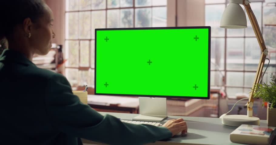 Professional Black Female Using Green Screen Monitor for Content Creation. Creative Woman working on Chroma Key Display. Modern Home Office with Warm Wooden Minimalist Interior. Over the Shoulder Shot
