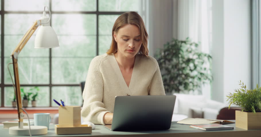 Portrait of Beautiful White Woman Working on Laptop at a Sunlit Desk. Room Has, Books, Plants and Large Windows, She Focuses on Digital Design, Social Media Marketing Campaign. Medium Portrait