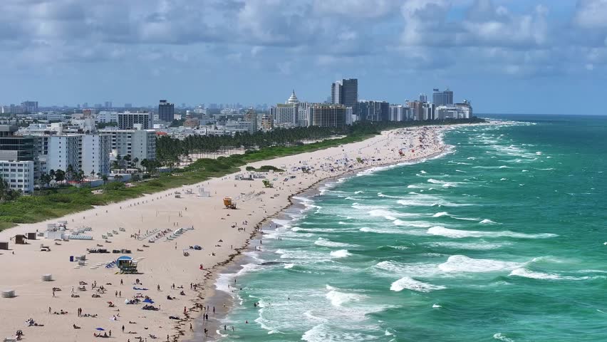Expansive view of South Beach’s coastline with sunbathers along the sandy shore. The Atlantic Ocean waves meet the beach, while Miami Beach’s skyline is visible in the background. Aerial.