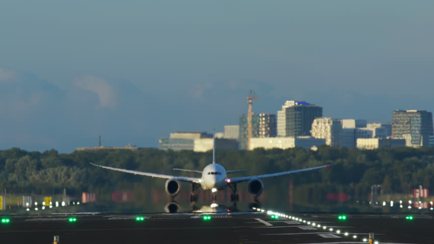 A large commercial airplane is seen taking off from a runway with landing lights on. The aircraft is captured frontally with its wheels lifted off the ground, set against a backdrop of distant
