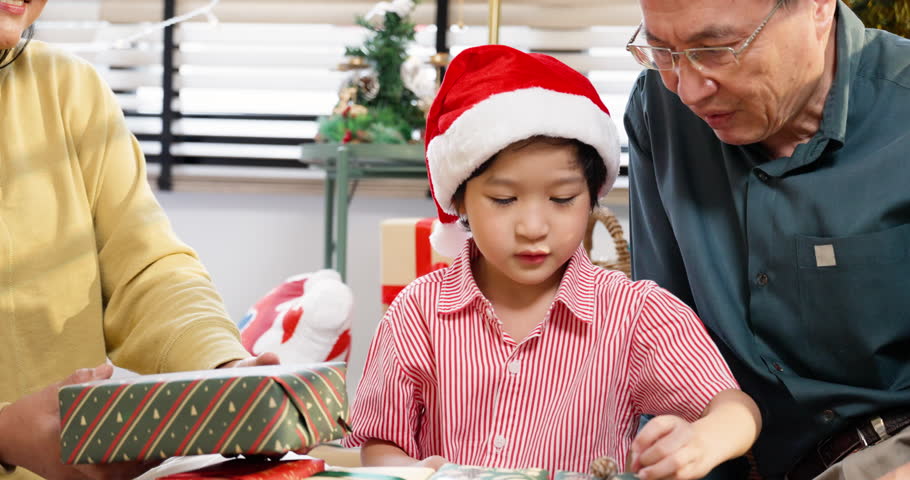 young Asian boy wearing Santa hat, carefully handling a wrapped Christmas present. Grandfather and middle-aged woman watching with warm smiles, love and anticipation during Christmas gift exchange.