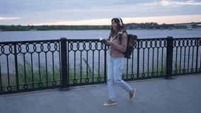 Young woman with headphones and a backpack enjoys reading a book while walking along a riverbank at sunset, embracing the tranquility of nature and the beauty of the evening sky - Powered by Shutterstock - Get 15% off with code: PIKWIZARD15
