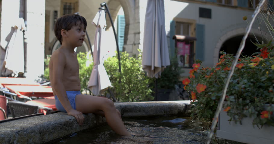 Young boy splashing water with his foot in a stone fountain, playful and carefree summer fun outdoors, in 800 fps slow motion, surrounded by colorful flowers