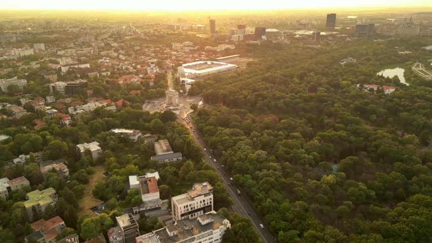 High angle drone footage of Herastrau and Arcul de Triumf in Bucharest Romania. Green park shot on a sunny day during golden hour. Cityscape of warm summer evening. Rugby stadium visible from above