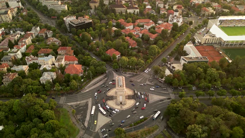High angle drone footage of Herastrau and Arcul de Triumf in Bucharest Romania. Green park shot on a sunny day during golden hour. Cityscape of warm summer evening. Rugby stadium visible from above