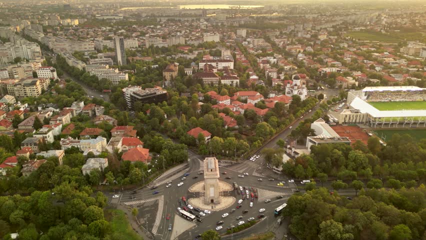 High angle drone footage of Herastrau and Arcul de Triumf in Bucharest Romania. Green park shot on a sunny day during golden hour. Cityscape of warm summer evening. Rugby stadium visible from above
