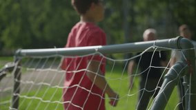 A soccer ball is shot across a goal post as the goalkeeper in red attempts to save it, while two people watch in the blurred background - Powered by Shutterstock - Get 15% off with code: PIKWIZARD15