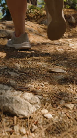 Vertical video. A Woman Climbing Up a Rocky Path with a View of the Sea in the Distance. Her Sneakers and Legs are in Focus, Emphasizing the Effort and Adventure of Hiking. The Sunlight Filters Throug