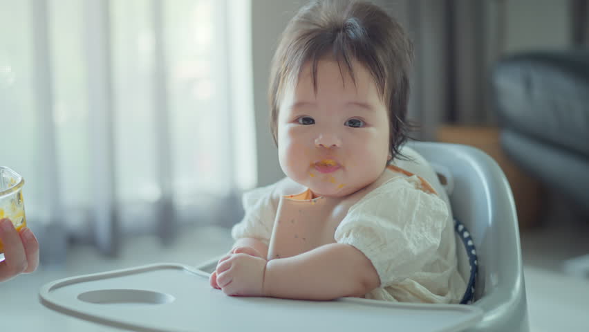 Closeup of Happy asian baby girl eating first food pumpkin from spoon with mom at home, Smile baby looking at camera.