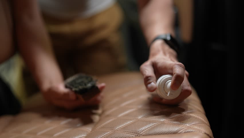 Close-up cropped shot of unrecognizable auto service man carefully cleaning car seat with brush and foam, focusing on detail to maintain vehicle. Brown leather upholstery being meticulously cleaned.