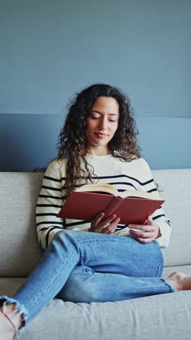 Young woman immersed in a captivating book while relaxing on a cozy couch. Young woman sitting on a couch, immersed in a captivating book, enjoying a peaceful moment of relaxation and leisure