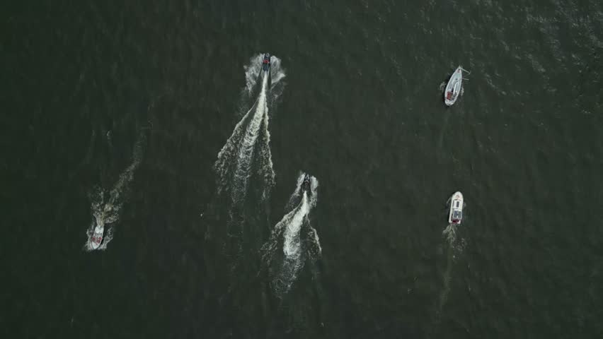 Boats creating wakes as they move across dark water in a calm, organized pattern, aerial view