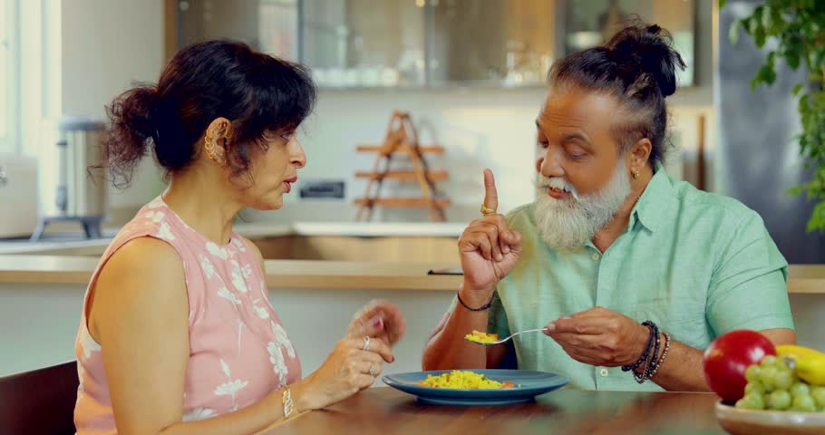 Indian Asian middle aged couple sitting at dining table,wife tasting food with spoon cooked by husband
