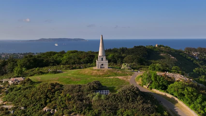Killiney Hill, County Dublin, Ireland, June 2023. Drone swiftly orbits clockwise around The Obelisk with panoramic views of Killiney Bay and Dublin City Center in the distance on a bright sunny day.