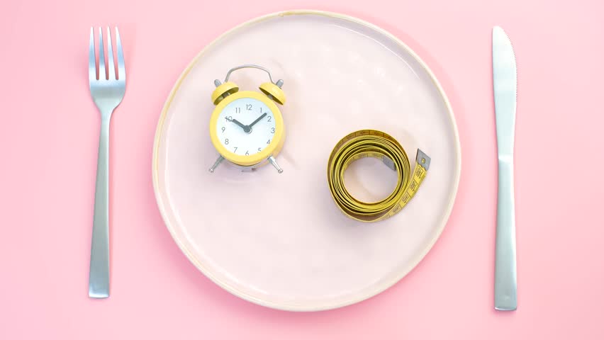 Empty plate with alarm clock and measuring tape on pink background, intermittent fasting concept.