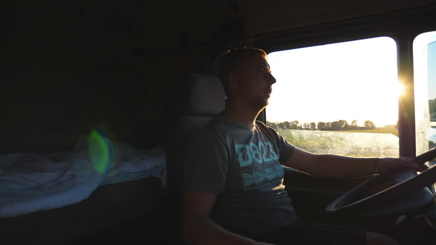 Man driving a truck and carefully watching the road. Caucasian guy is riding through the countryside on the sunset background. Profile of lorry driver inside the cab. Side view Slow motion Close up