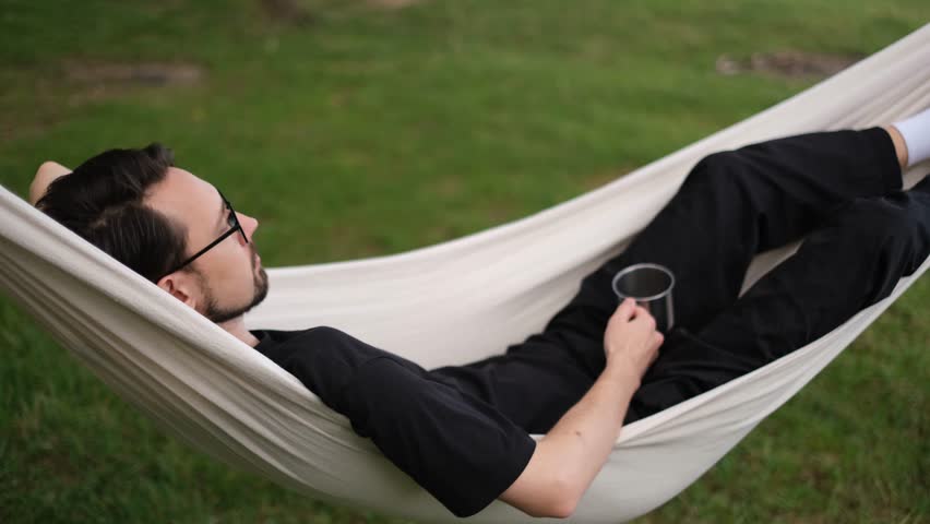  young man relaxing in a hammock and drinking tea in the forest by the sea on a sunny day