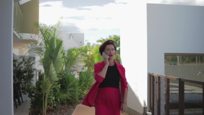 Businesswoman in vibrant red suit walk while chatting on phone. Smiling brightly, Girl enjoy lively conversation