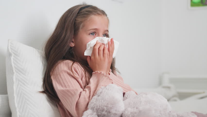 Sick girl sneezing snot into tissue paper in bedroom at home. Female child blowing runny nose is suffering from cold and flu 