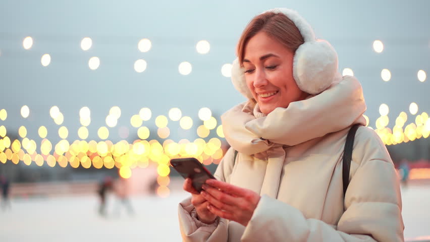 Smiling woman in winterwear using smartphone while standing at Christmas market. Happy female is looking at camera outdoors during festive season 