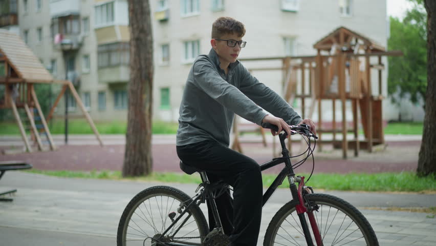 Boy in a grey top and black trousers sits on his bicycle in a park setting, he is resting while holding the bike handlebars, the background features a playground structure and apartment buildings
