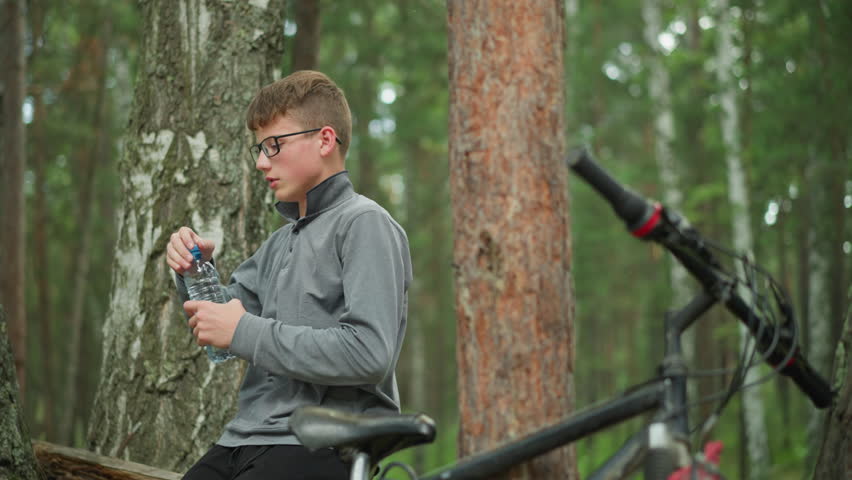 Young boy wearing glasses and grey long-sleeve top drinks from a water bottle, taking a break in a forest surrounded by tall green trees, a bicycle is parked beside him in the background