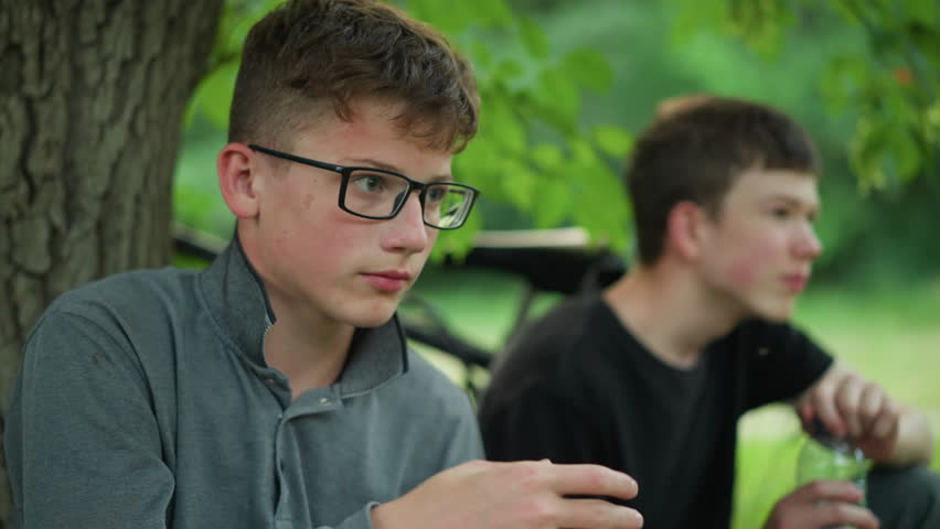 Two young siblings resting under a tree with one of them drinking water while the other bows his head, they are taking a break from a bike ride, with bicycles parked nearby surrounded by greenery