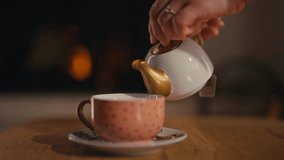 Close-up of a female hand pouring hot tea from a teapot into a cup, creating a cozy ambiance with a warm fireplace in the background. Concept of relaxation and warmth - Powered by Shutterstock - Get 15% off with code: PIKWIZARD15