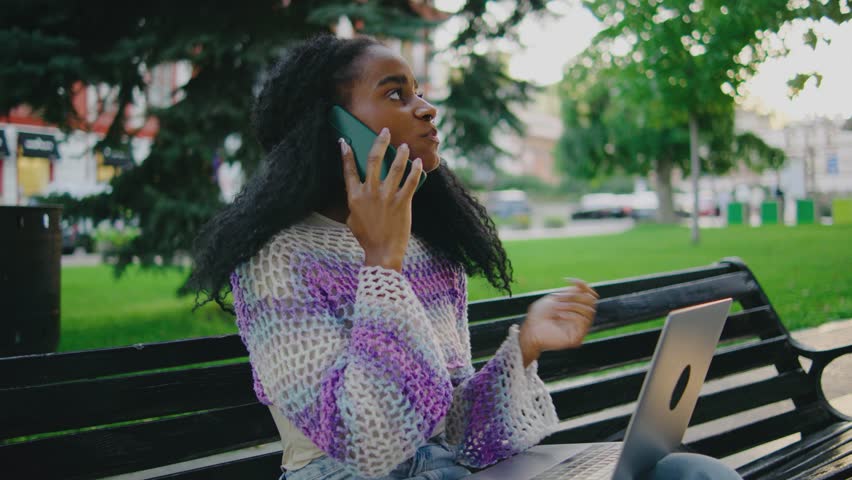 A young woman is actively engaged in a phone call while diligently working on her laptop, all in a serene park setting, surrounded by the beauty of nature and vibrant greenery