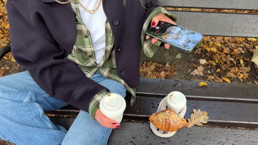 A female blogger is making a photo of coffee and a croissant against the background of autumn leaves in the park while sitting on a bench