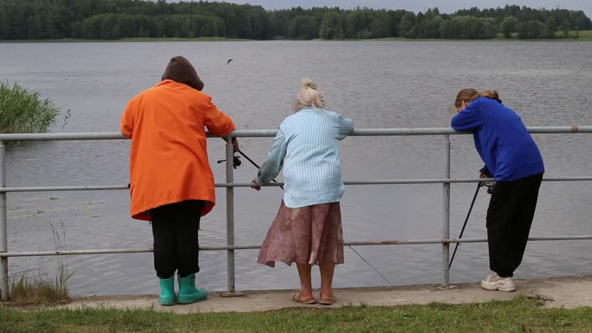 Family outdoor activities - daughter, mom and grandmother fishing on lake in windy weather. Two fisherwomen and girl with fishing rod angling on river in small village, green forest in the background