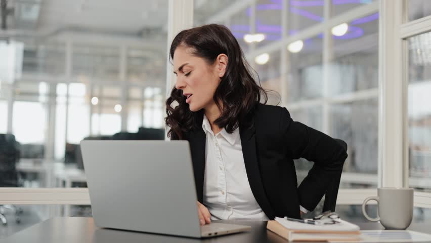Young business woman suffering from back pain while working on laptop in office. Neck pain, backache, spinal problem. Tired employee after long work. Exhausted female executive massaging lower back.
