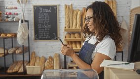 Woman in a bakery shop using a smartphone, surrounded by bread and pastries, with menu board in background, wearing apron and glasses, mature female managing business indoors - Powered by Shutterstock - Get 15% off with code: PIKWIZARD15