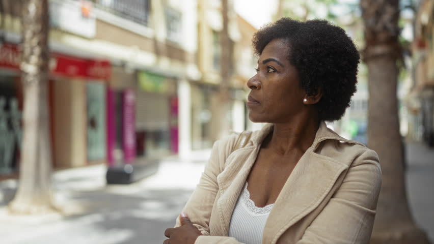 Woman standing confidently with arms crossed in an urban setting displaying african american curly hair and an elegant beige jacket